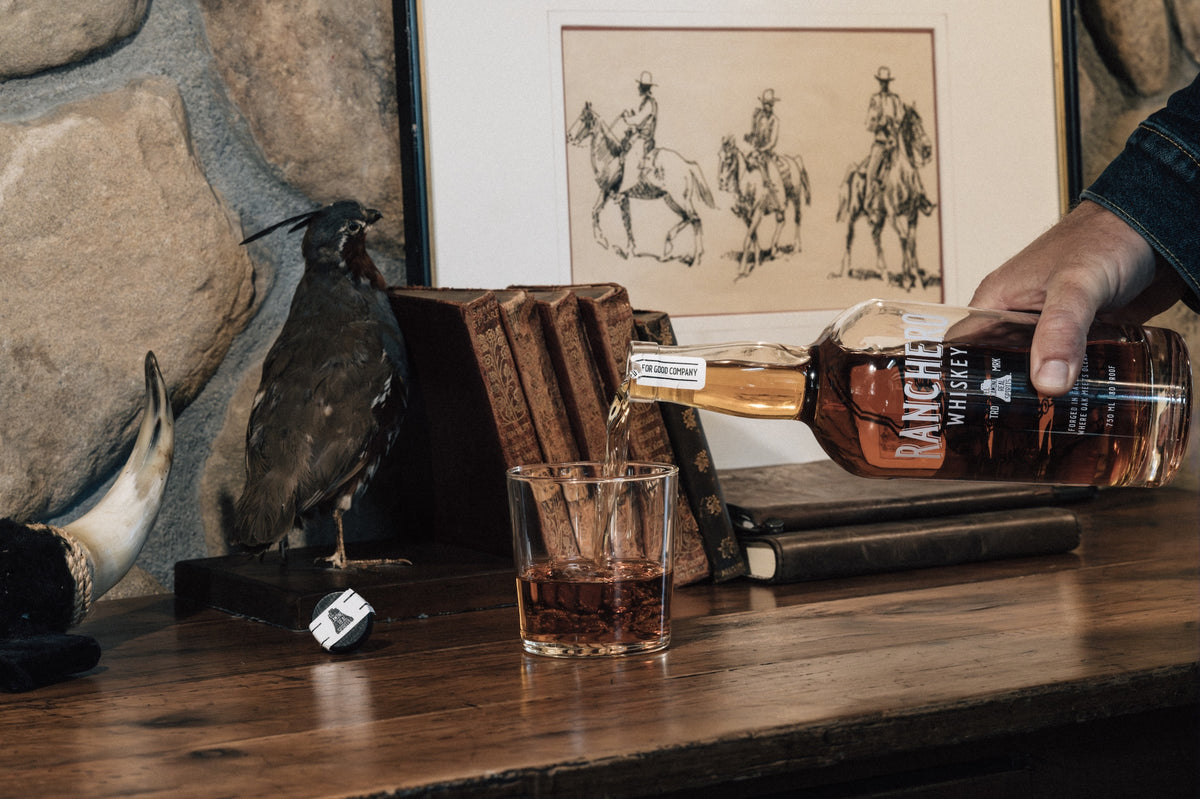 A hand pours amber liquor from a bottle labeled Ranchero into a clear rocks glass on a wooden table, with framed artwork, books, and a taxidermy bird in the background.